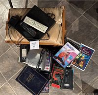 Wide overhead photo showing all items in the lot including DVD player, remotes, VHS tapes, CD/alarm clock, book, and foot rest glider