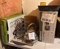 Photo showing glass tea kettle with metal handle and lid, metal spice rack with multiple glass jars, stacked wicker charger plates, boxed electric wok, and a boxed roasting pan visible.