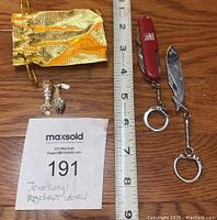 All items laid out including two fish pendants, two keychains (one fish-shaped), red pocket multi-tool, and gold drawstring pouch next to measuring tape for scale.