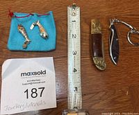 Photo showing two vintage silver fish pendants with turquoise eyes next to a blue pounch, an antique gold tone folding pocket knife with wooden handle, a black fish utensil keychain, and a measuring tape for scale.