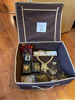 View of open fabric box with assorted jewelry including brooches, necklaces, bracelets, a watch, and earrings.