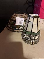 Two stained glass lamp shades, one smaller squared shade in white and green and one larger round shade with floral pattern.