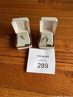 Two 14 KT gold necklace charms each in its own small jewelry box on wood floor with MaxSold lot ticket.