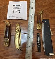 All five pocket utensils laid flat near a measuring tape to show size, including Thai gold tone folding knives and black utensils.