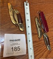 All four pocket utensils and multi-tools on wooden surface alongside a measuring tape showing items about 4 to 5 inches long and a MaxSold lot card 185