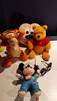 Four Disney character plush toys: Tigger, a large Mickey Mouse face cushion, Winnie the Pooh wearing red shirt, Mickey Mouse baby plush in blue pajamas with bib