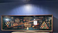 Full view of shadow box displayed on a blue wall, showing all elements inside including billiard balls in racks, cue, and historical plaques.