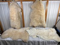 Four cream to off-white wool floor mats displayed on and hanging from a wooden frame, showing thick, fluffy wool texture and irregular natural shapes.