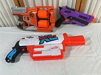 Three Nerf dart guns on a white surface, including an orange double drum revolver, a purple Fortnite pistol, and a white and orange Mega XL gun.