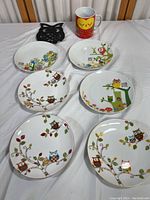 Six owl-themed plates, one owl mug, and one black owl trivet arranged on a table. Plates feature colorful owl illustrations on white background.