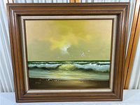 Framed ocean landscape painting showing sea waves, cloudy sky, seagulls, and sandy beach with rocks.