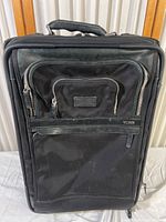 Front view of black Tumi rolling suitcase showing multiple zippered compartments and textured fabric with some wear marks.