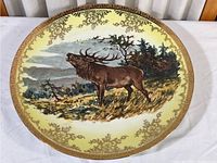 Top view of the vintage elk charger plate showing detailed painted elk and surrounding nature scene with gold gilded floral border