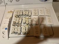 Top-down photo showing multiple white duplex receptacles and several matching wall plates arranged neatly on a surface.