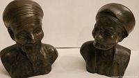 Two bronze bust sculptures of a man and woman, positioned side by side on a white surface showing details of head coverings and facial features.