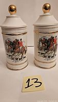 Front view of two white porcelain cookie jars with gold trim and lids topped with gold spheres. Each jar is decorated with detailed painted scene of man riding a horse with hunting dogs running alongside.