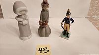Front view of 3 porcelain figurines: woman holding chicken, woman staying warm, English soldier