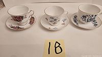 Three tea cup and saucer sets shown from side view displaying floral designs and gold rims.