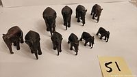 Set of 10 dark brown wooden elephant figurines shown from an elevated angled front view. The sizes range from large to small. Some tusks are visibly broken on a few elephants.