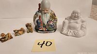 Five Buddha and Asian figure statues displayed on a flat surface with a label '40'. Two adult porcelain Buddhas and three small wooden carvings visible.