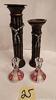 Two tall metal candle holders with rope design and two small red and clear glass candle holders with floral etching placed on a white surface with a number card.