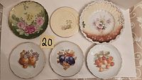 Full view of 6 decorative plates with various floral and fruit designs, some with gold trim, arranged on a table.