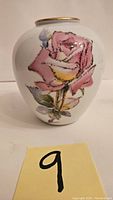 Front side of white porcelain vase with hand-painted large pink rose with green leaves and yellow center, gold trim on top rim.