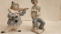 Two Tengra porcelain clown figurines side by side; one playing violin, the other holding a toy instrument.