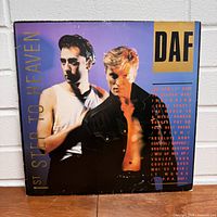 Front cover of DAF 1st Step to Heaven vinyl album. Shows two men, background gradient purple to black, gold and red text with track titles.