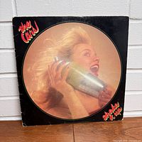 Photo of the front cover of The Cars 'Shake It Up' vinyl record album featuring an image of a woman holding a cocktail shaker, with the band's name and album title in red and yellow text.