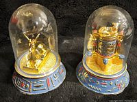 Two Franklin Mint Egyptian figurines with golden figures under clear glass domes on blue bases decorated with hieroglyphics. Missing bottom covers on the bases visible.