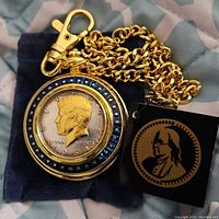 Gold-tone Franklin Mint pocket watch featuring 1964 JFK coin face and attached chain, laid on blue velvet cloth with original tag included.