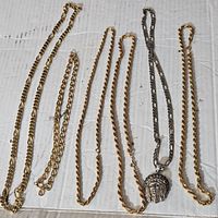 Seven chains laid out on white surface, gold-tone and silver-tone metals visible, one chain with Jesus Christ head pendant