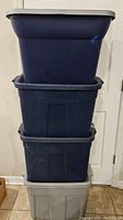 Stack of four plastic storage totes, three navy blue and one light gray, all with lids, stacked against a wall.