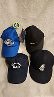 Four baseball caps laid out on beige textile background showing front logos and designs: blue Corona Extra, black Nike, black Greatest Dad, black with white bear graphic.