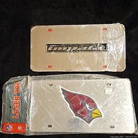Two license plates, one with 'Impact' text and the other with NFL Cardinals logo, both in original plastic packaging, viewed from front