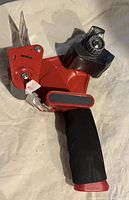 Heavy duty tape dispenser with red metal body, black cushioned handle, and metal blade for cutting tape, shown on a light background.