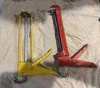Pair of two manual caulking guns lying flat on a white cloth surface. One gun is yellow, the other is red, both showing visible wear and usage marks.