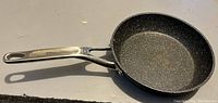 Photo showing a round frying pan with a speckled non-stick inner cooking surface, metal handle marked with 'THE ROCK'.