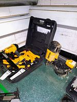 Two Bostitch nail machines displayed on a workbench. One stored in open black carrying case with collated yellow plastic nails and lubricant bottle visible. Second is a coil siding nailer with coil nails loaded.