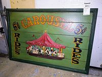 Front view of a rectangular carved wooden sign painted green with a 3D pink-roofed carousel in the center, and text stating "5¢ CAROUSEL 5¢ RIDES RIDES" in yellow and black letters.