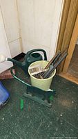Close-up photo of the boot scraper and items in the bucket including metal rods and sockets, with watering can in background.