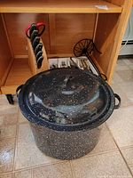 Photo showing large speckled black enamel pot with lid positioned in front of the wooden kitchen cabinet shelf holding cutlery block, flatware organizer tray, and paper towel holder.