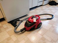 Red Dirt Devil Easy Lite canister vacuum with hose and wand placed on tiled floor near kitchen appliances.