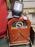Front view of red Lincoln Electric AC-225 amp arc welder with black helmet and gloves on top