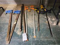 Photo showing a row of seven clamps on a floor, varying in size and material, some with blue handles, orange handles, and black/yellow triggers.