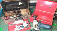 Wide view showing Milwaukee Screwshooter in its original metal case with accessories and the Bosch jigsaw in the red metal carrying box, laid out on a workbench.