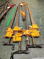 Four Pony metal bar clamps arranged side by side on a workshop floor, showing the full length of the steel bars and the orange clamp heads with black screw handles.