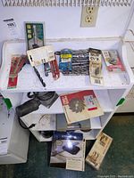 Full view of shelf with assorted drill bits laid out, red double bar clamp, PorterCable sander on middle shelf, Delta drum sander kit box, Rockler jig box, hardware packages.