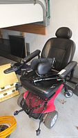 Full side and front view of the Pride electric powerchair showing red base, black cushioned seat, armrests, wheels and attachments